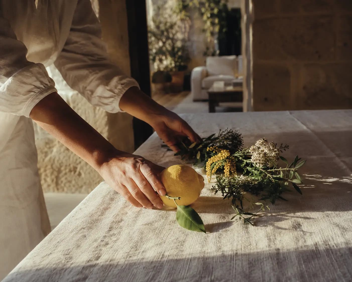 Mani che posano un limone e fiori freschi su un tavolo di benvenuto ricoperto di lino, in un appartamento siciliano in pietra